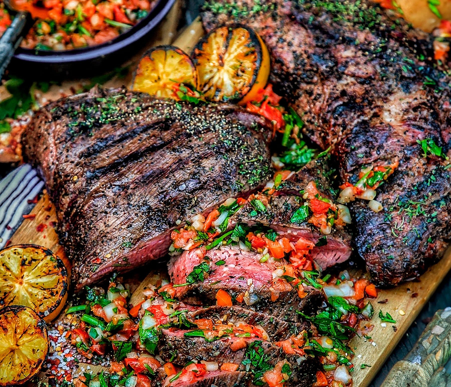 Grilled Santa Maria-style tri-tip beef sliced on a wooden board, garnished with fresh herb salsa and grilled lemons.