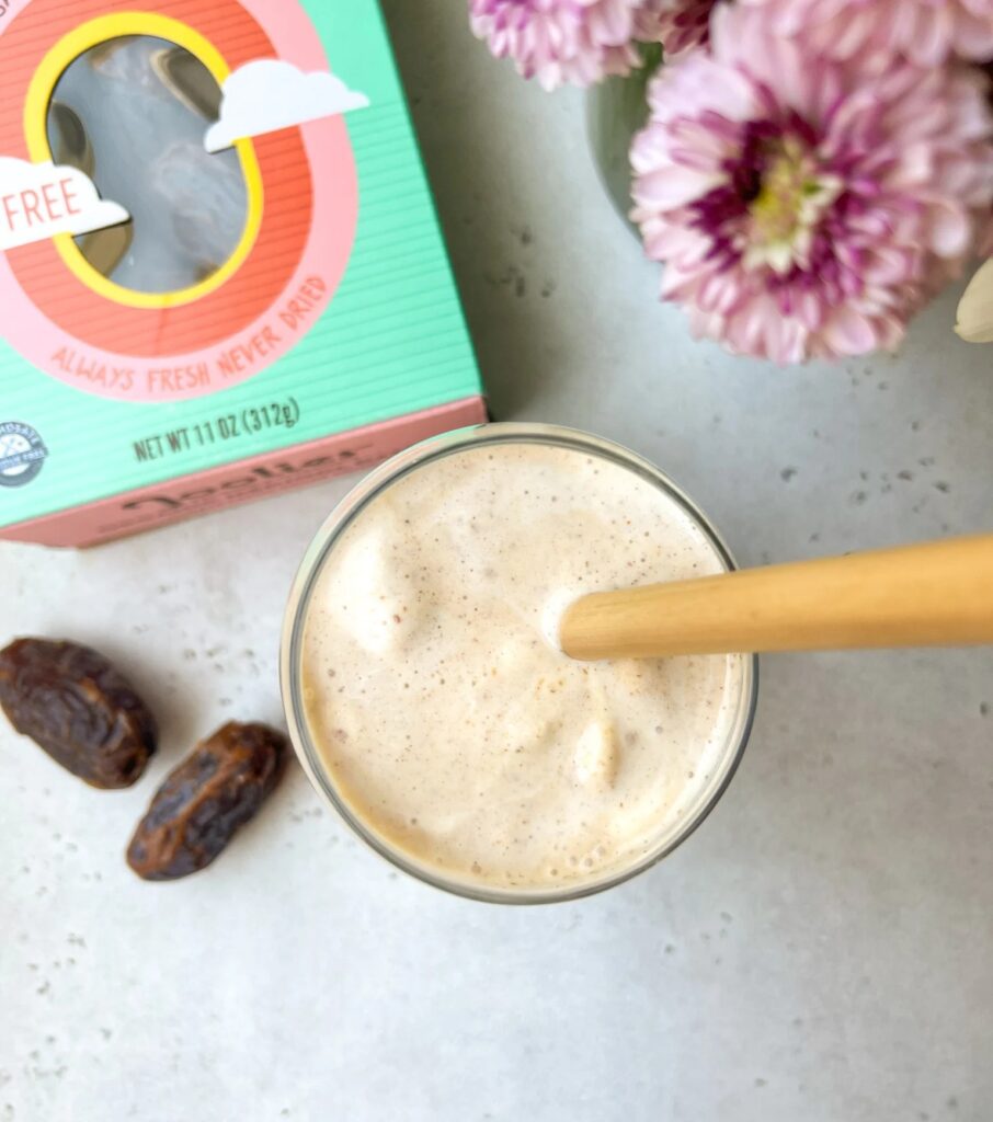 A creamy date shake in a glass with a bamboo straw, next to fresh Medjool dates and a colorful box.
